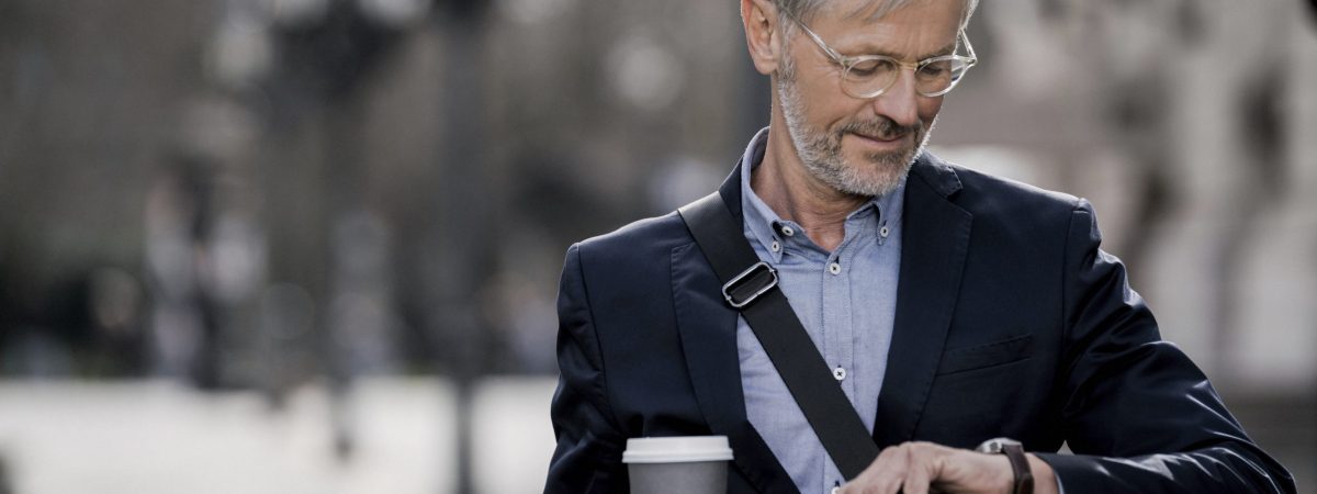 Grey-haired businessman in the city looking at watch holding coffee to go